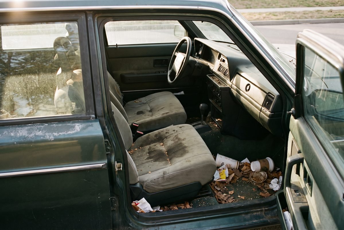 A neglected car interior with heavily soiled seats, multiple stains and debris in the footwells.