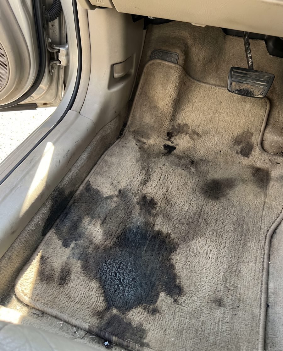 Car carpet with a large dark liquid stain soaked deep into the pile and the padding beneath.
